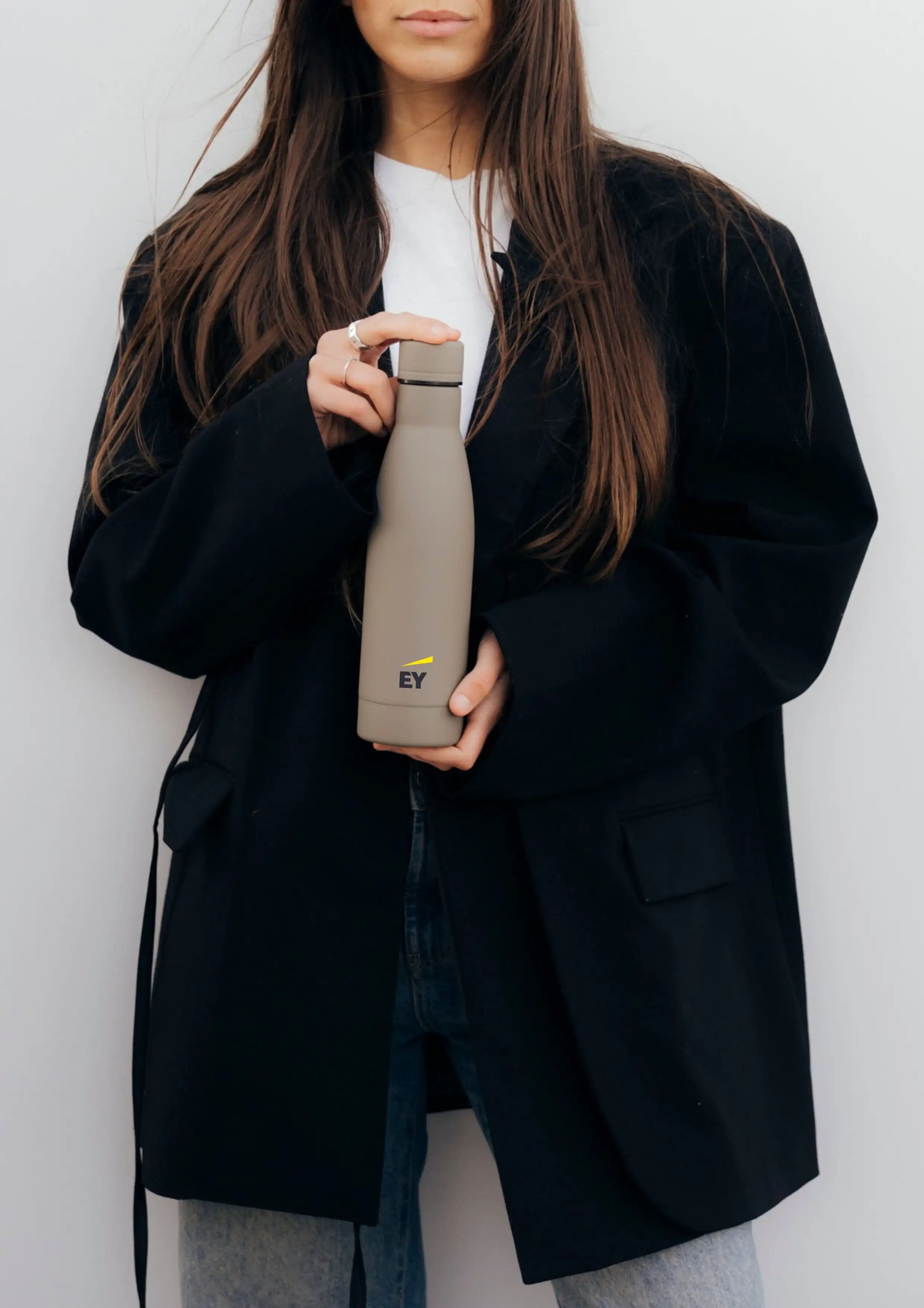 Botella térmica light brown Pilatus Brand con logotipo de EY sostenida por una mujer con chaqueta negra.