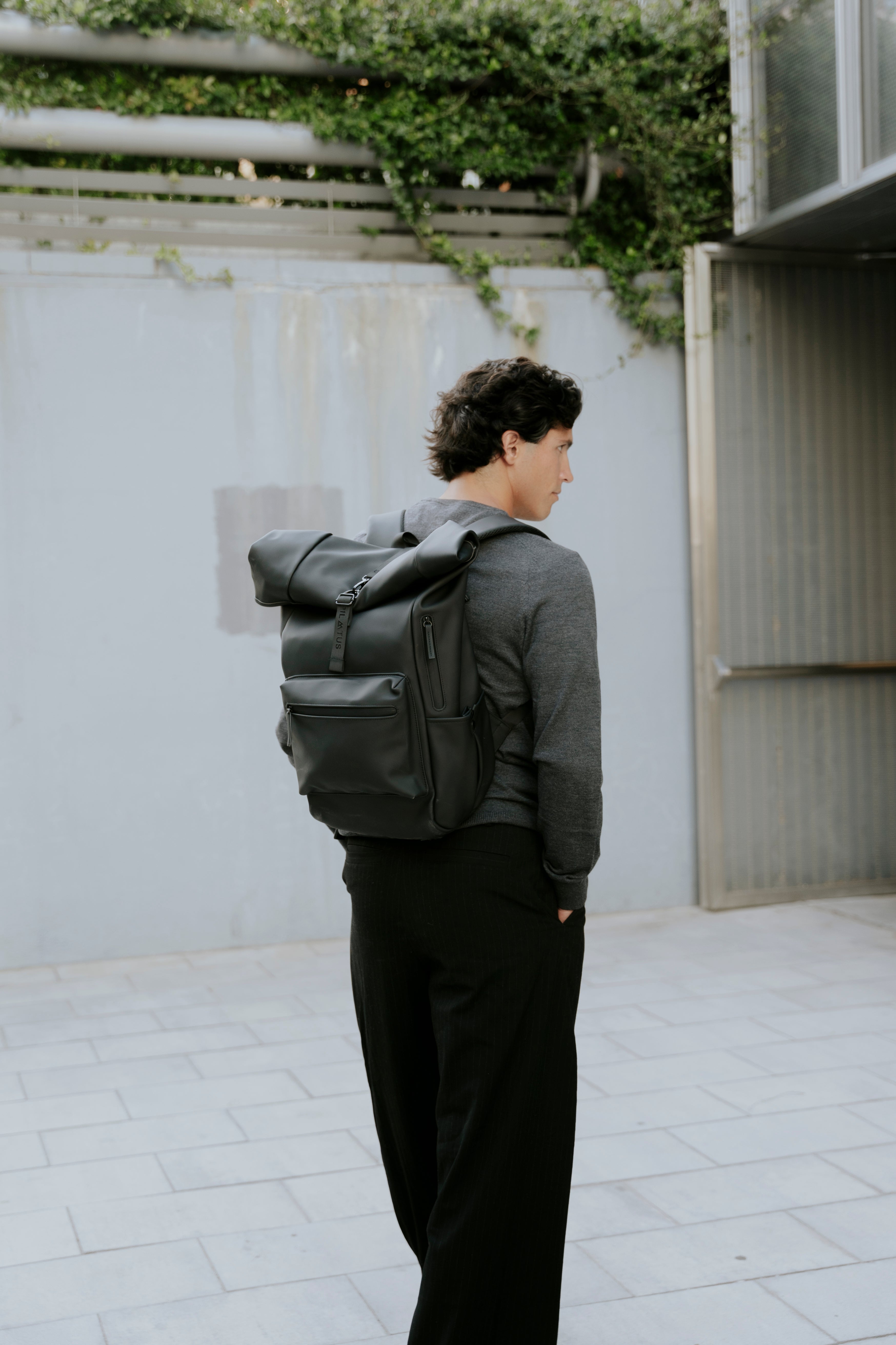 Mochila negra tipo roll-top de Pilatus Brand en la espalda de un hombre con jersey gris, exterior urbano