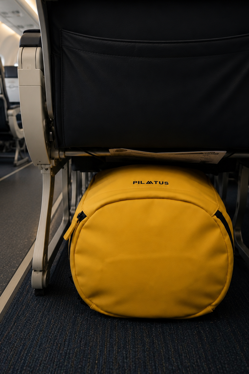Bolsa circular amarilla colocada bajo el asiento de un avión; alfombra azul y respaldo negro visibles. Texto: PILATUS