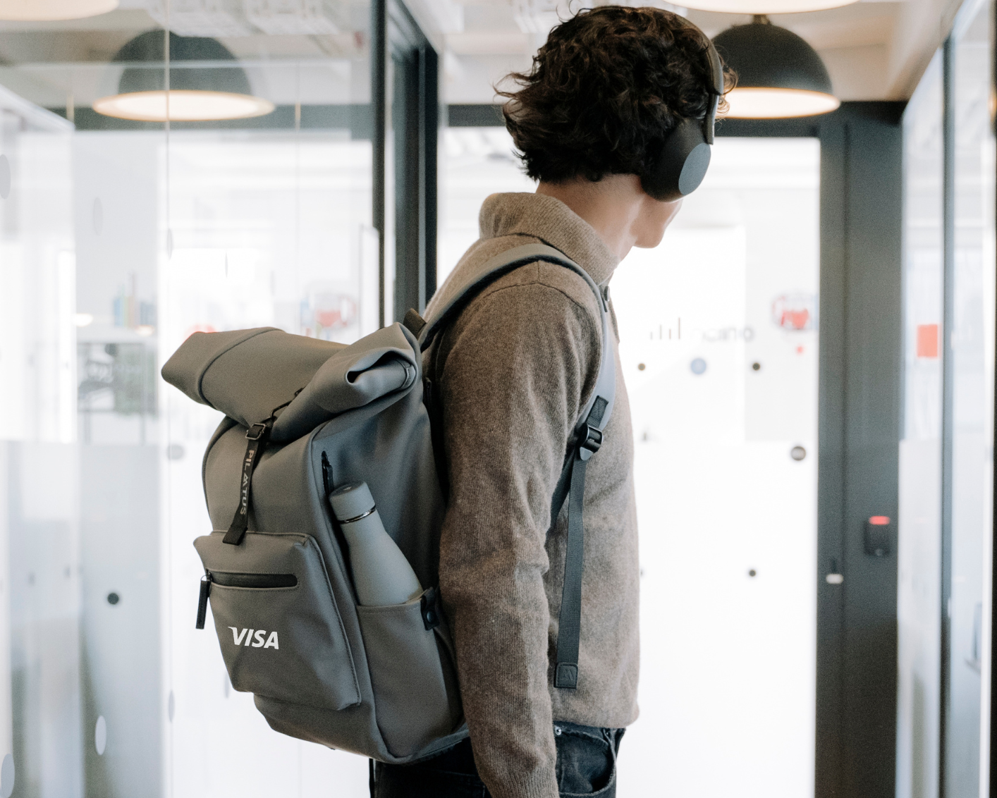 Hombre con auriculares llevando una mochila gris personalizada Pilatus Brand con logotipo de empresa VISA y botella reutilizable en el lateral