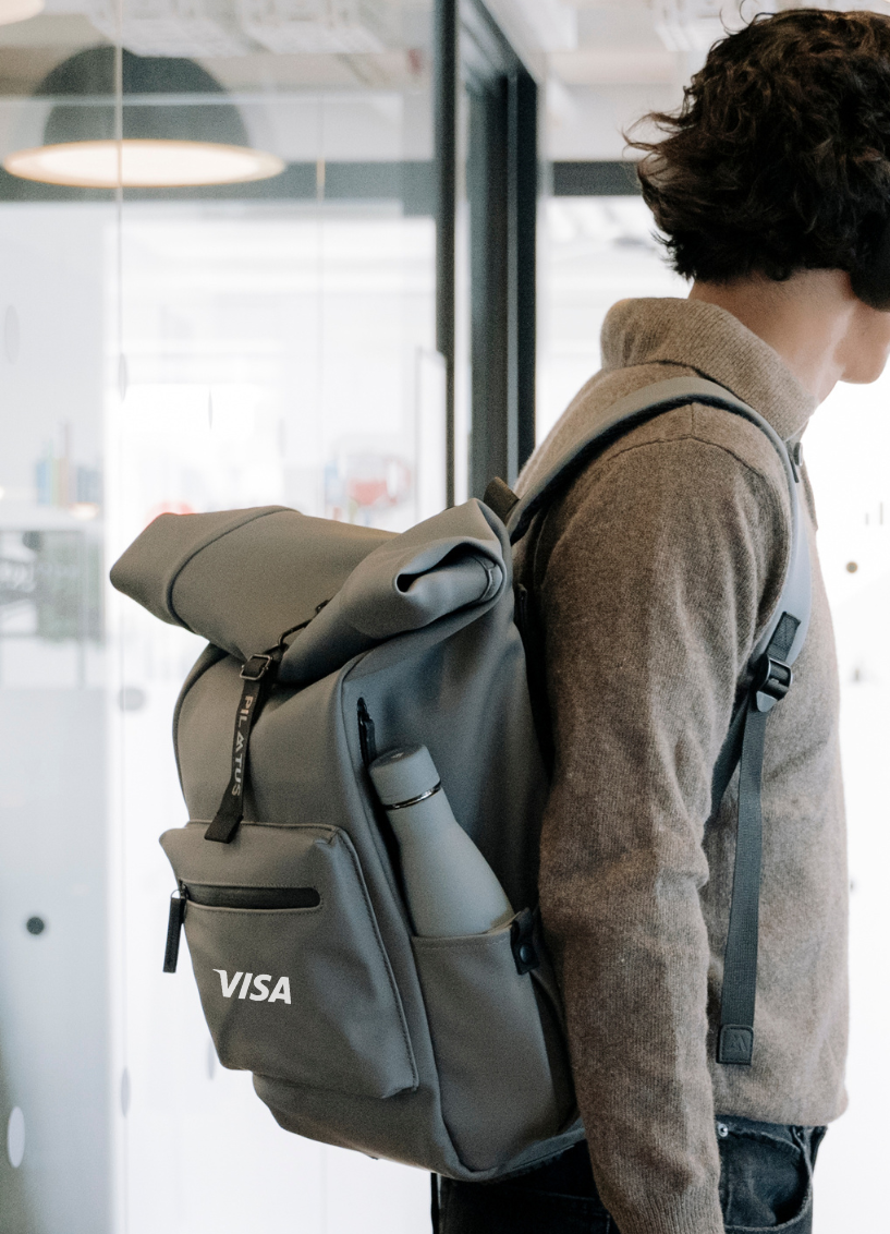 Hombre con auriculares llevando una mochila gris personalizada Pilatus Brand con logotipo de empresa VISA y botella reutilizable en el lateral