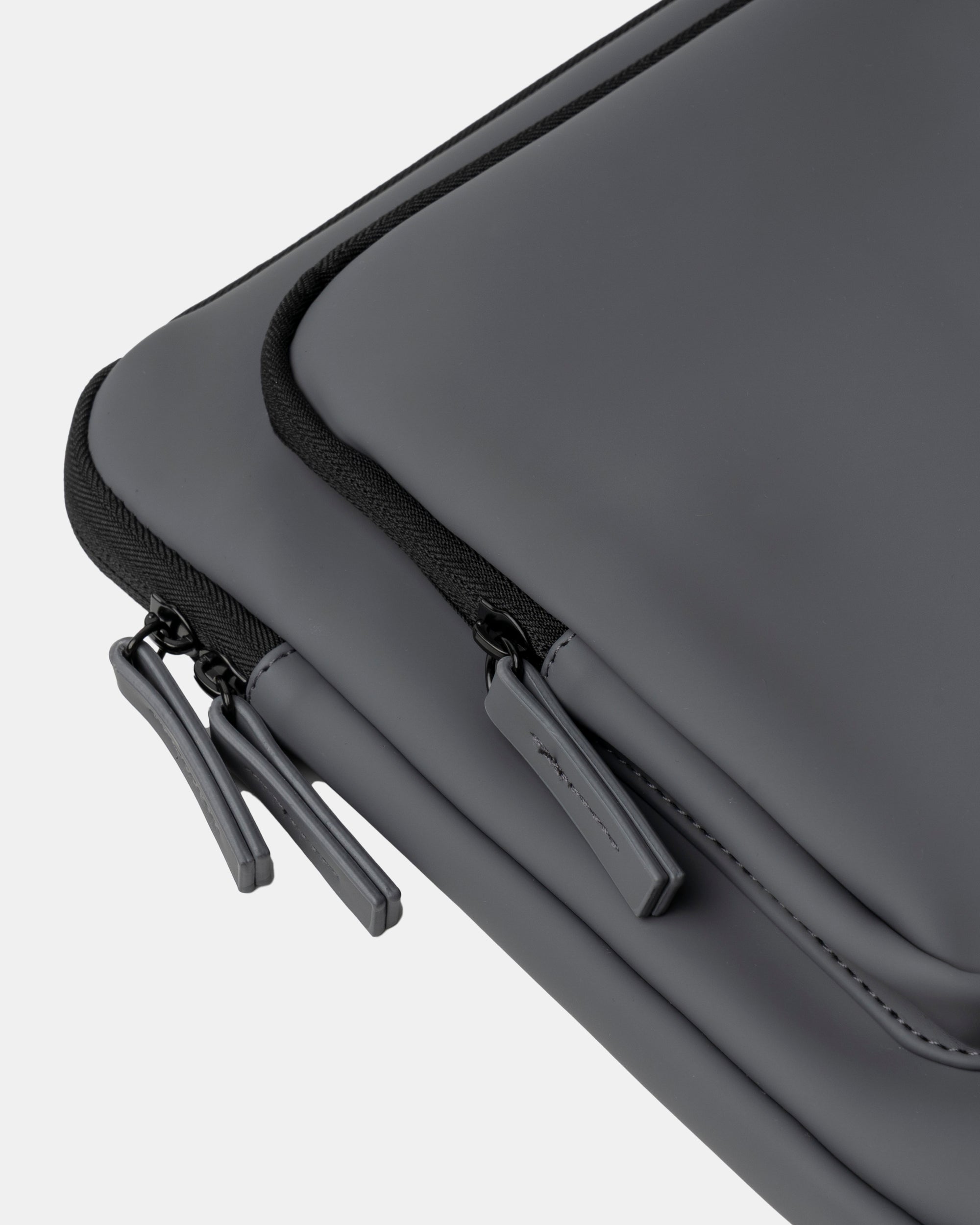 Mochila gris oscuro con exterior liso y cremalleras dobles, tiradores de goma y ribete negro; primer plano muestra costuras