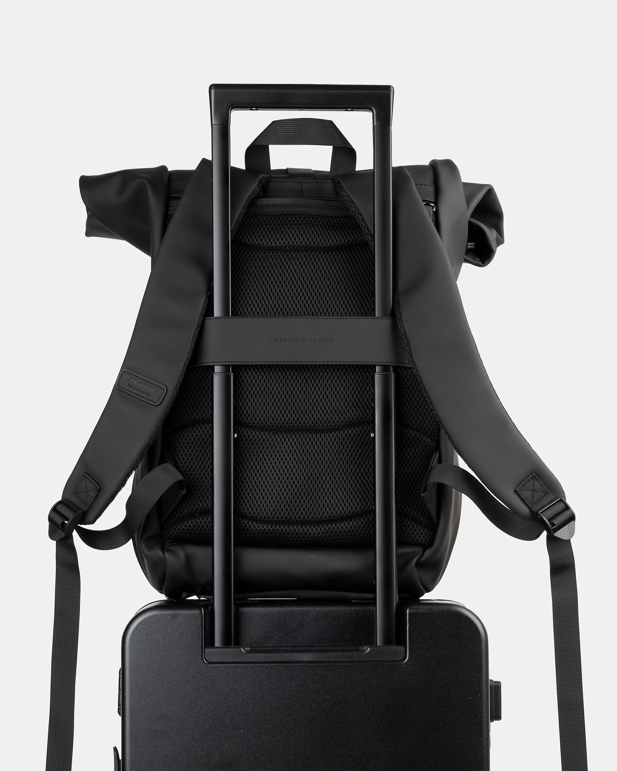 Mochila negra con respaldo acolchado y correas, montada sobre el tirador telescópico de una maleta. Texto: PILATUS (etiqueta)