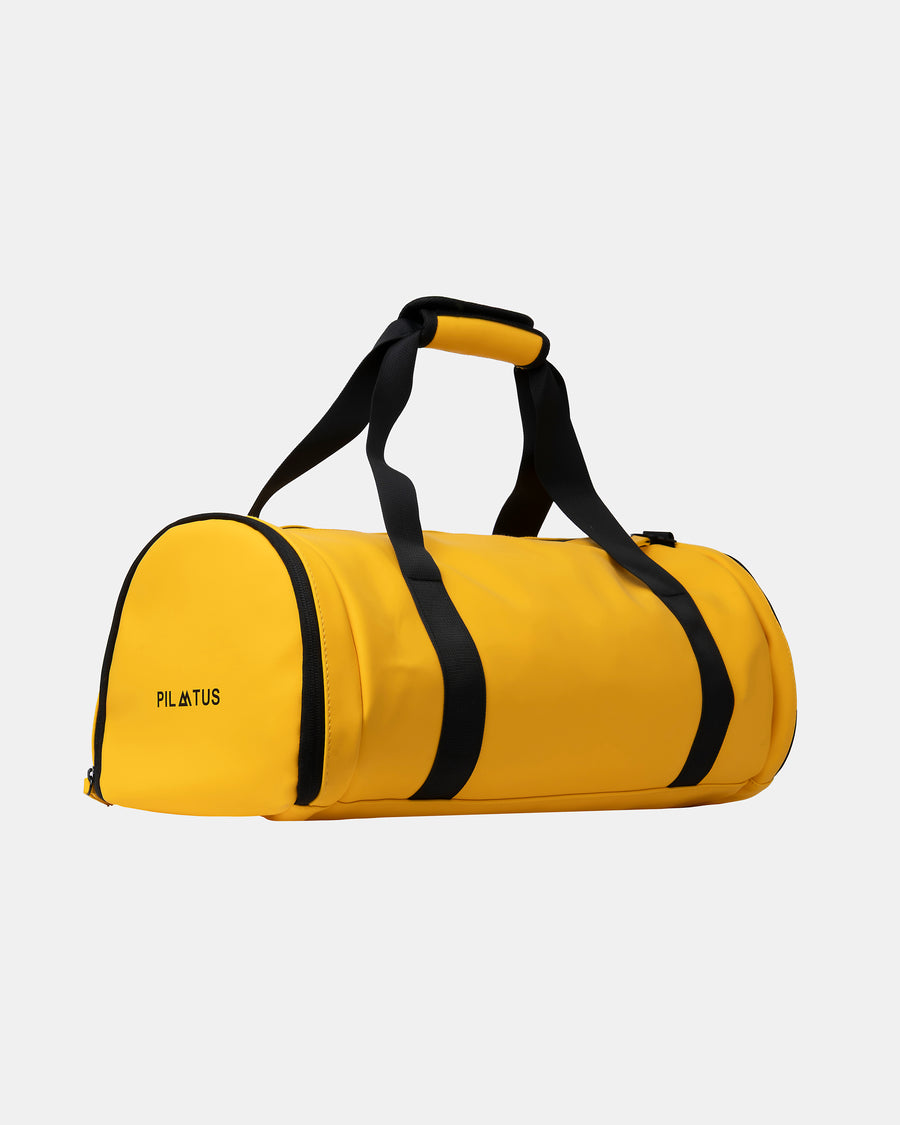 Bolsa de deporte amarilla con asas y correa negras, asa superior acolchada; fondo blanco neutro. Texto: PILATUS