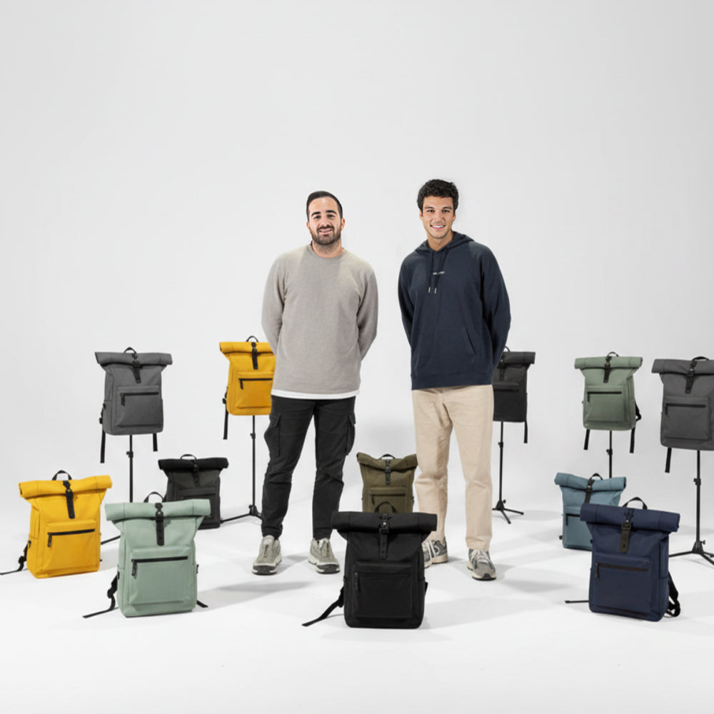 Mochilas enrollables de varios colores (amarillo, verde, azul, negro, gris) dispuestas en soportes y en el suelo