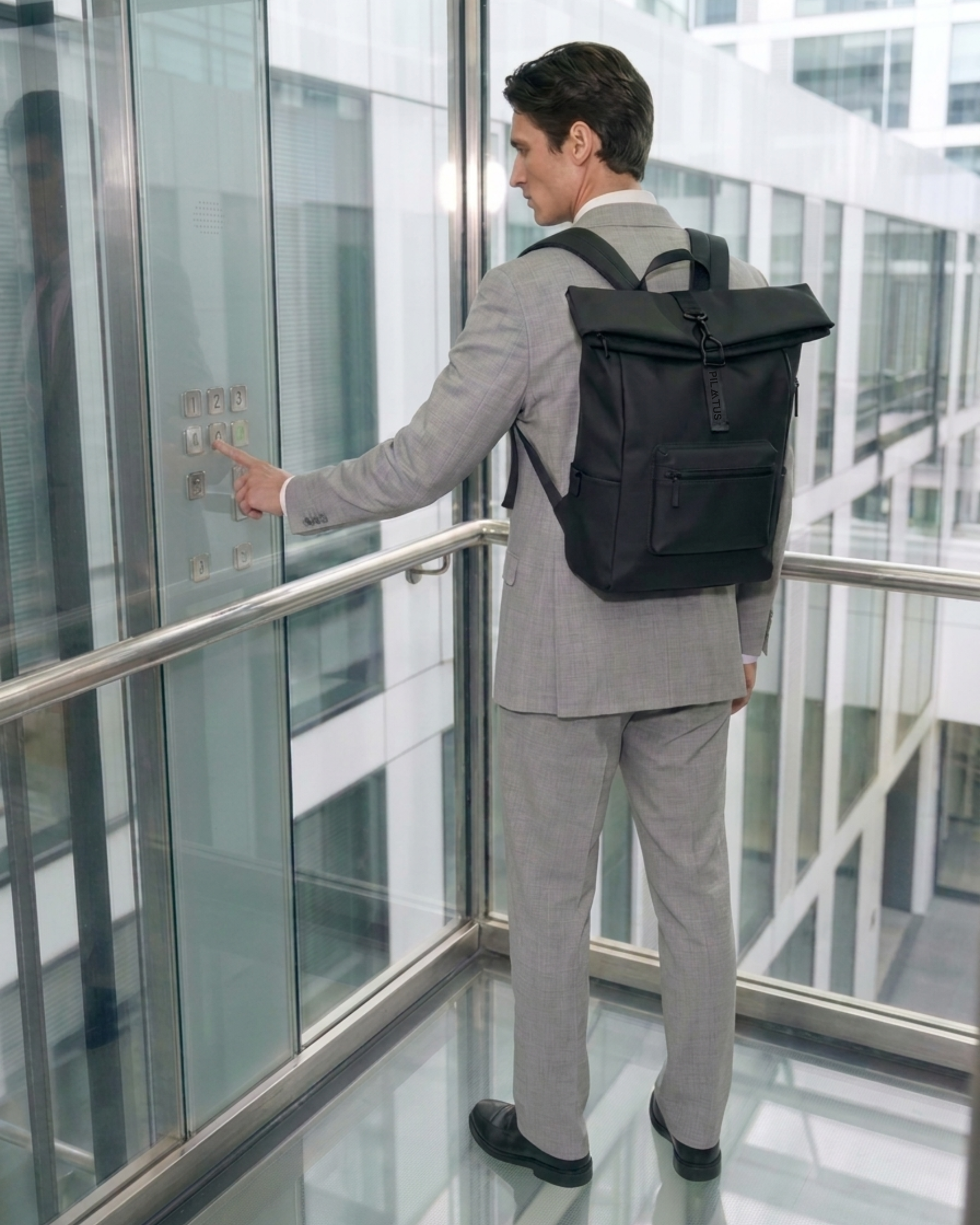 Mochila negra tipo roll-top sobre la espalda de un hombre con traje; en un ascensor de vidrio. Texto: PILATUS / 1 2 3