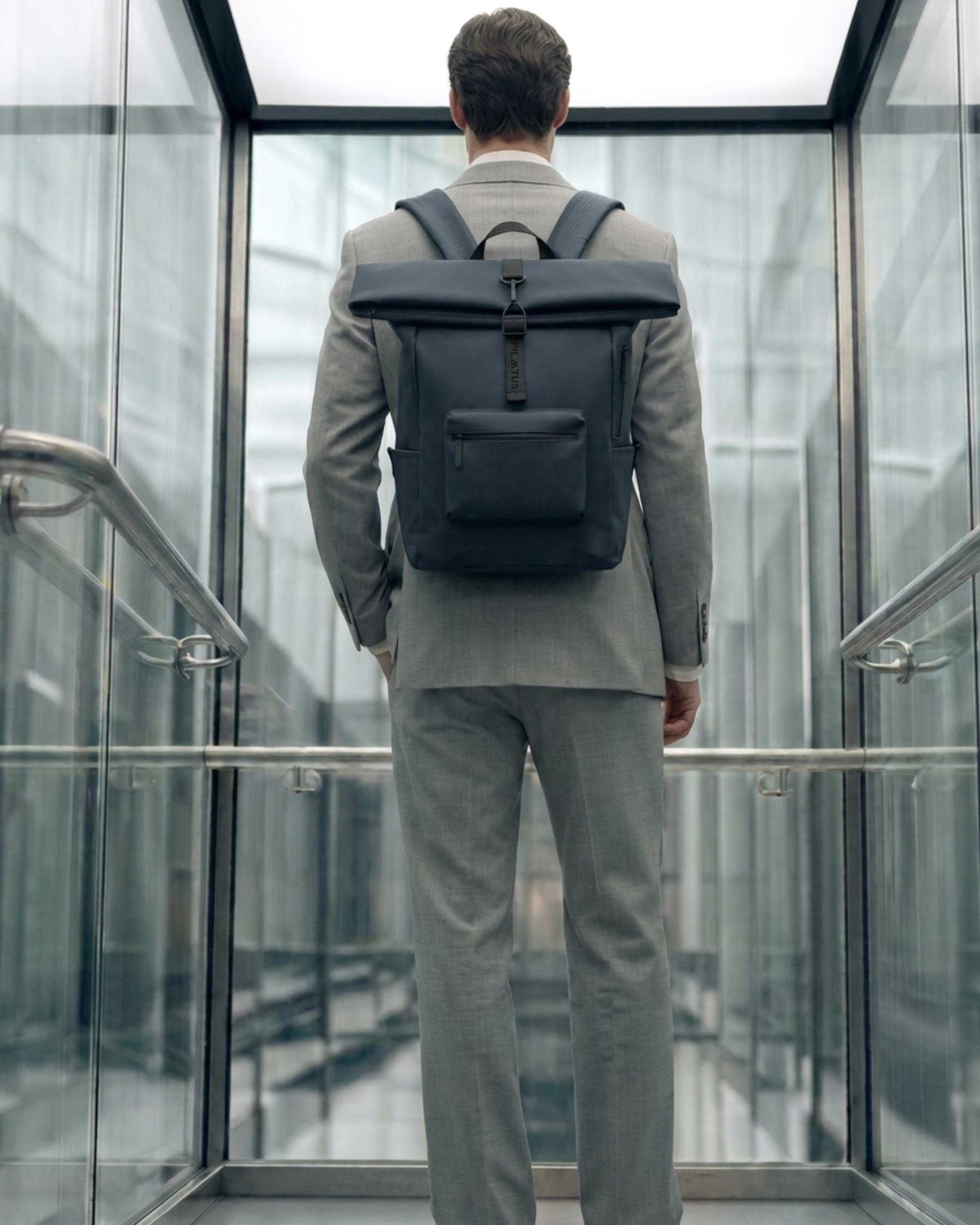 Mochila gris oscuro con cierre enrollable y bolsillo frontal; llevada por un hombre de traje gris. Texto: PILATUS