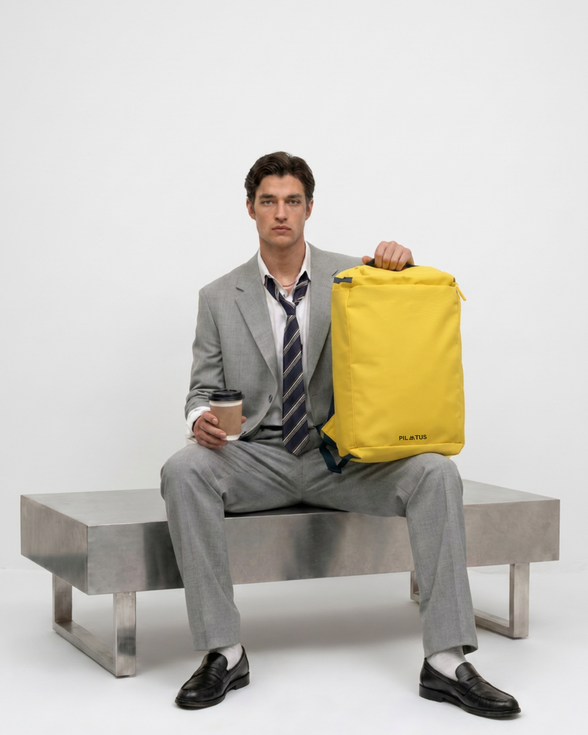 Mochila amarilla rectangular; hombre con traje gris sentado la sujeta; sostiene un vaso de café. Texto: PILATUS
