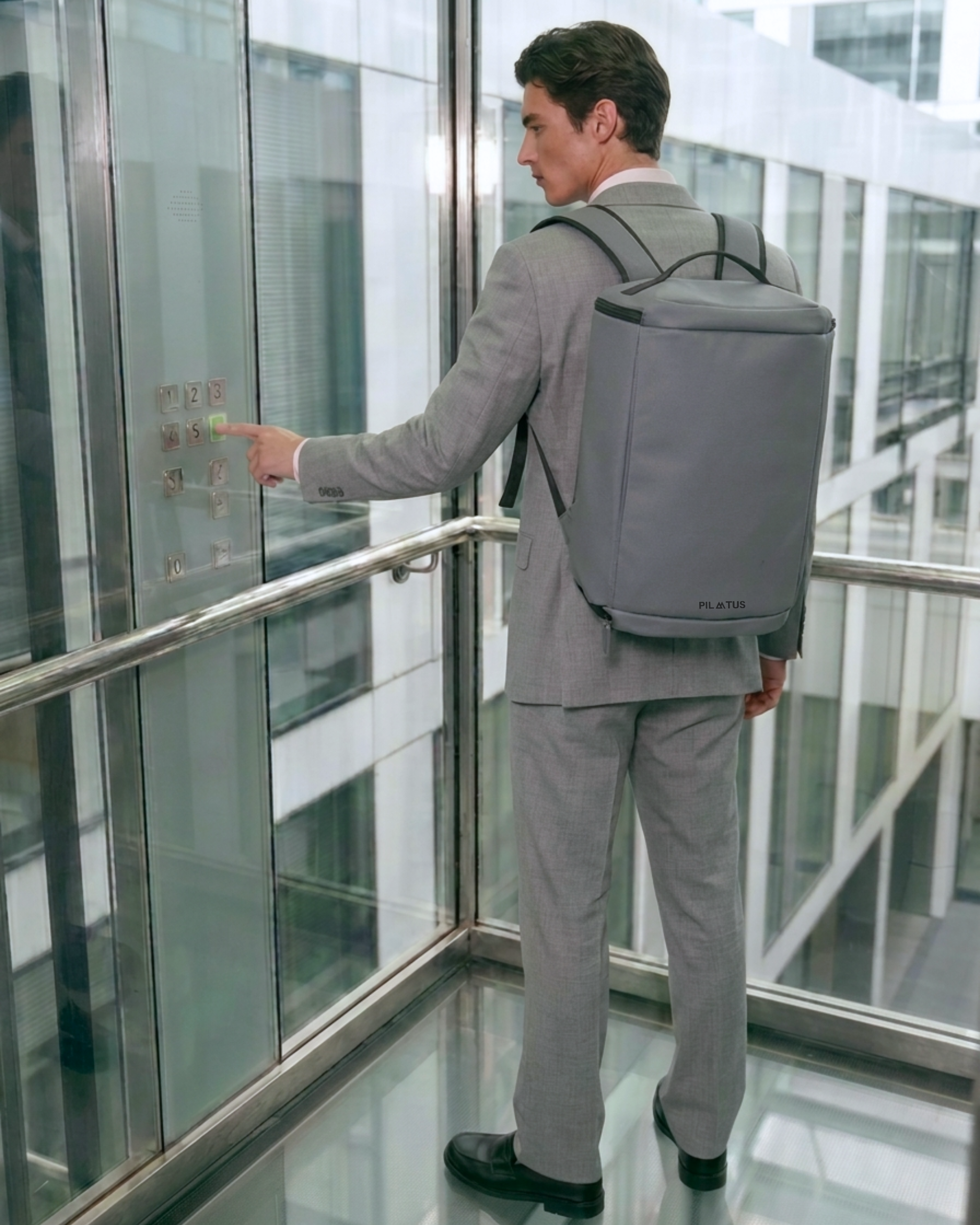 Mochila gris rectangular (marca: PILATUS) en la espalda de un hombre con traje; dentro de un ascensor de cristal
