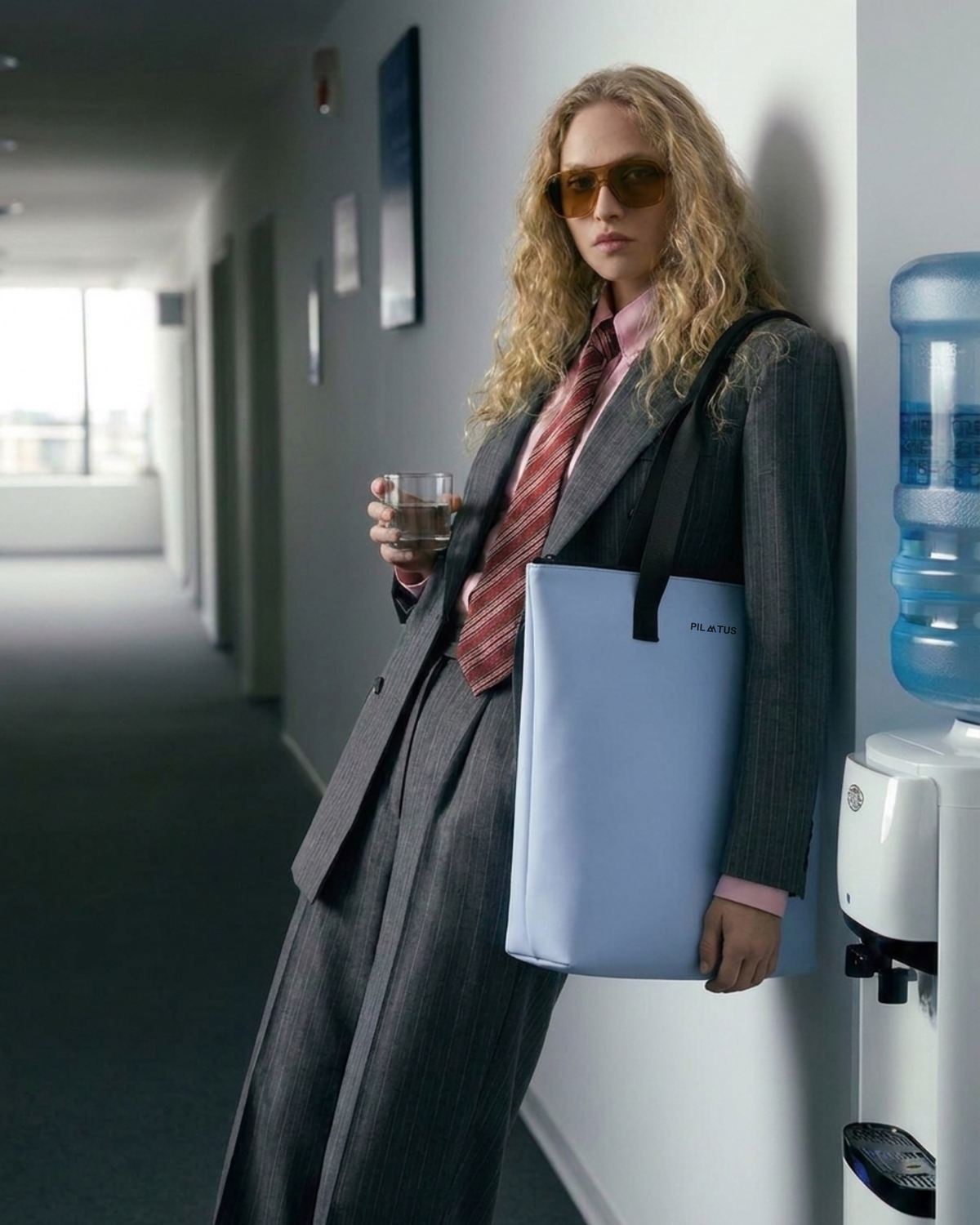 Bolsa tote celeste con el texto PILATUS, colgada del hombro de una persona con traje gris; sostiene un vaso