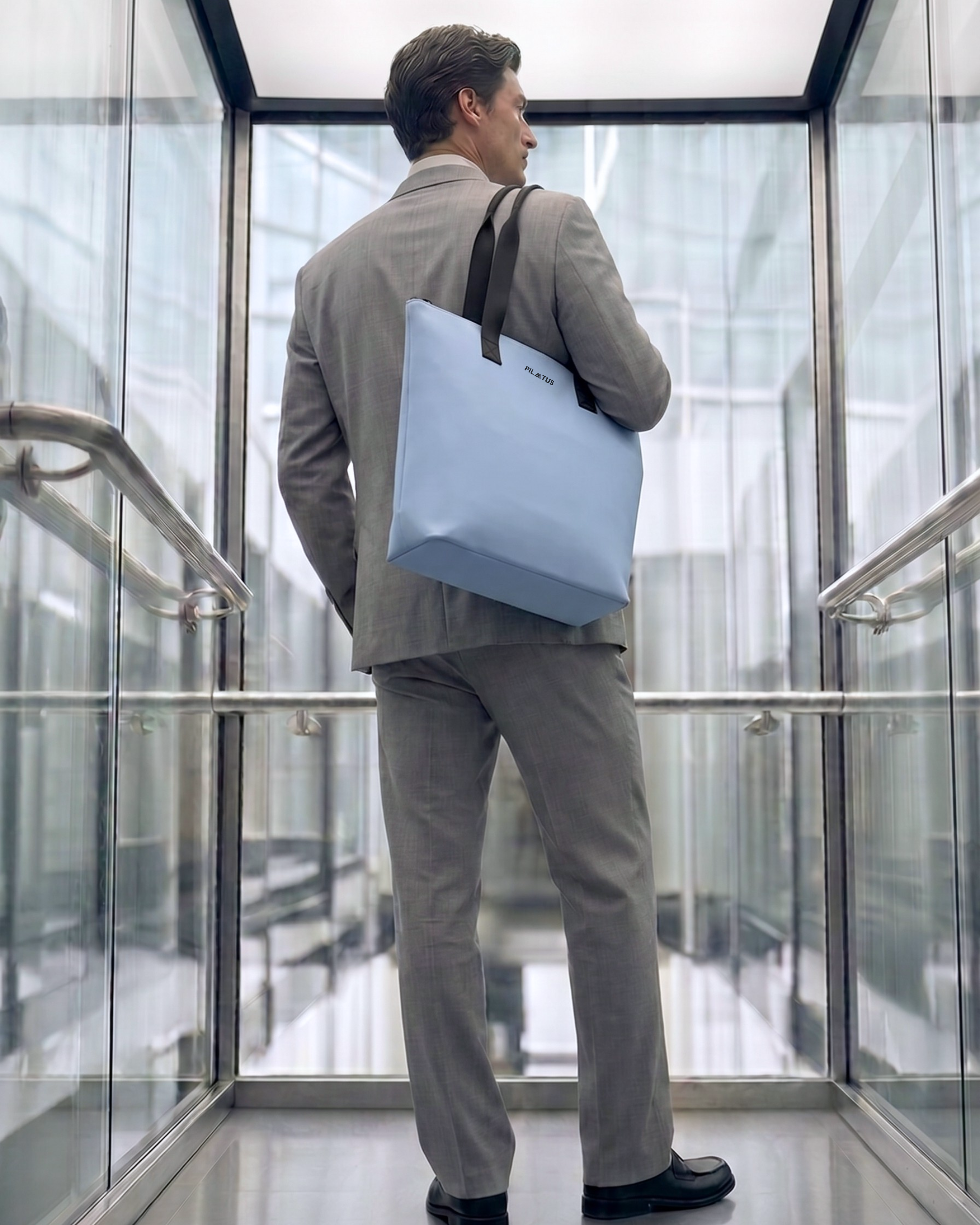 Bolsa tote azul claro con asas negras y texto PILATUS; hombro de un hombre con traje gris en ascensor de cristal
