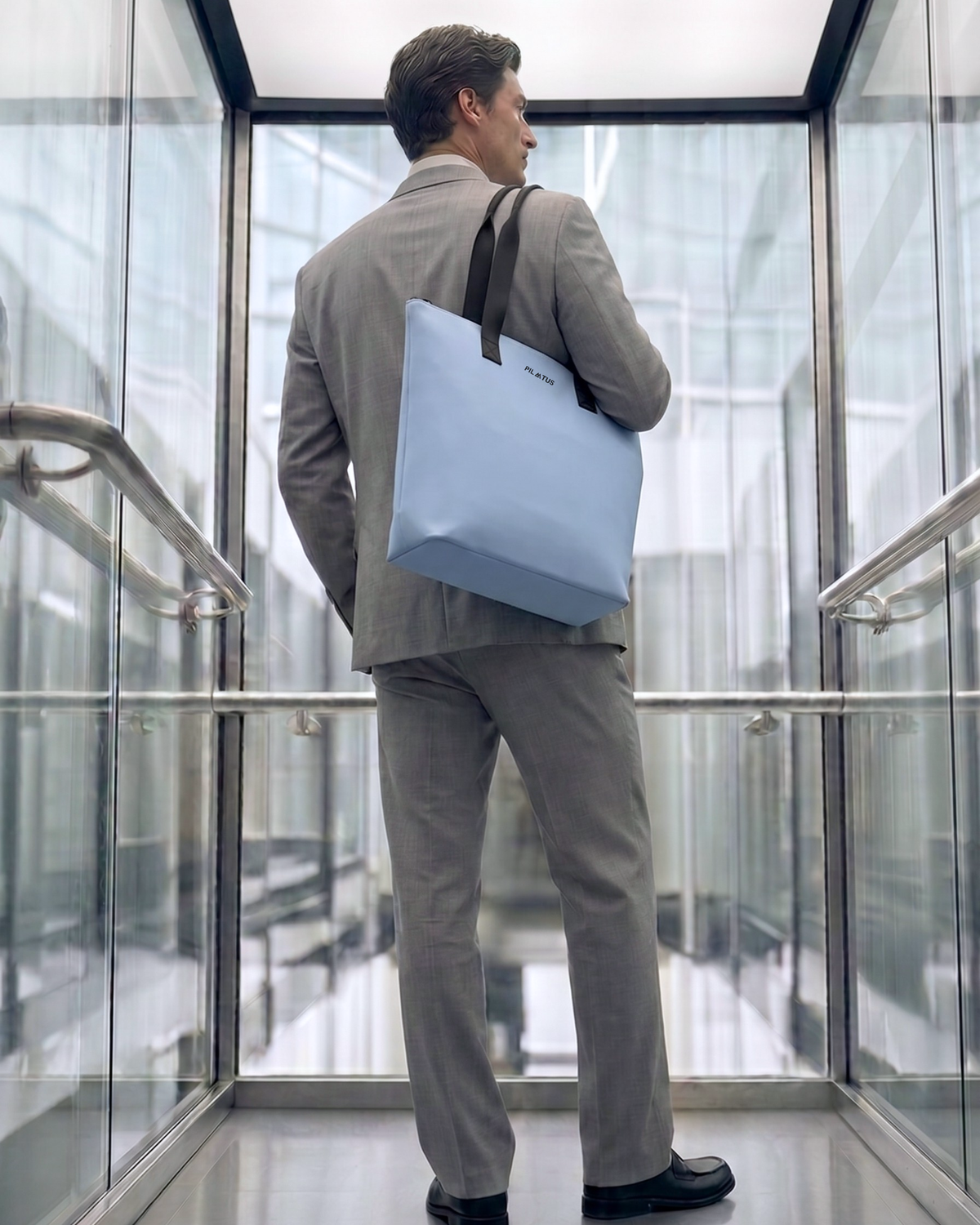 Bolsa tote azul claro con asas negras y texto PILATUS; hombro de un hombre con traje gris en ascensor de cristal