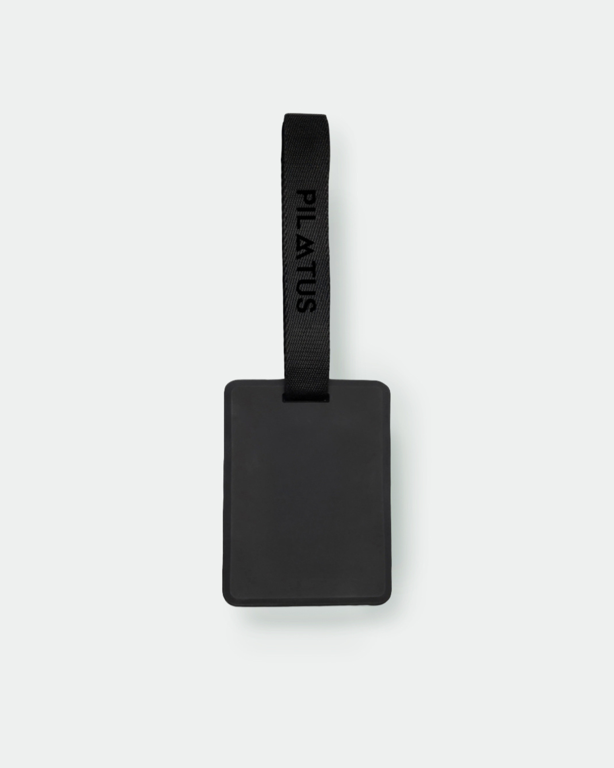 Etiqueta identificadora rectangular negra con correa textil negra marcada PILATUS; suspendida sobre fondo claro minimalista
