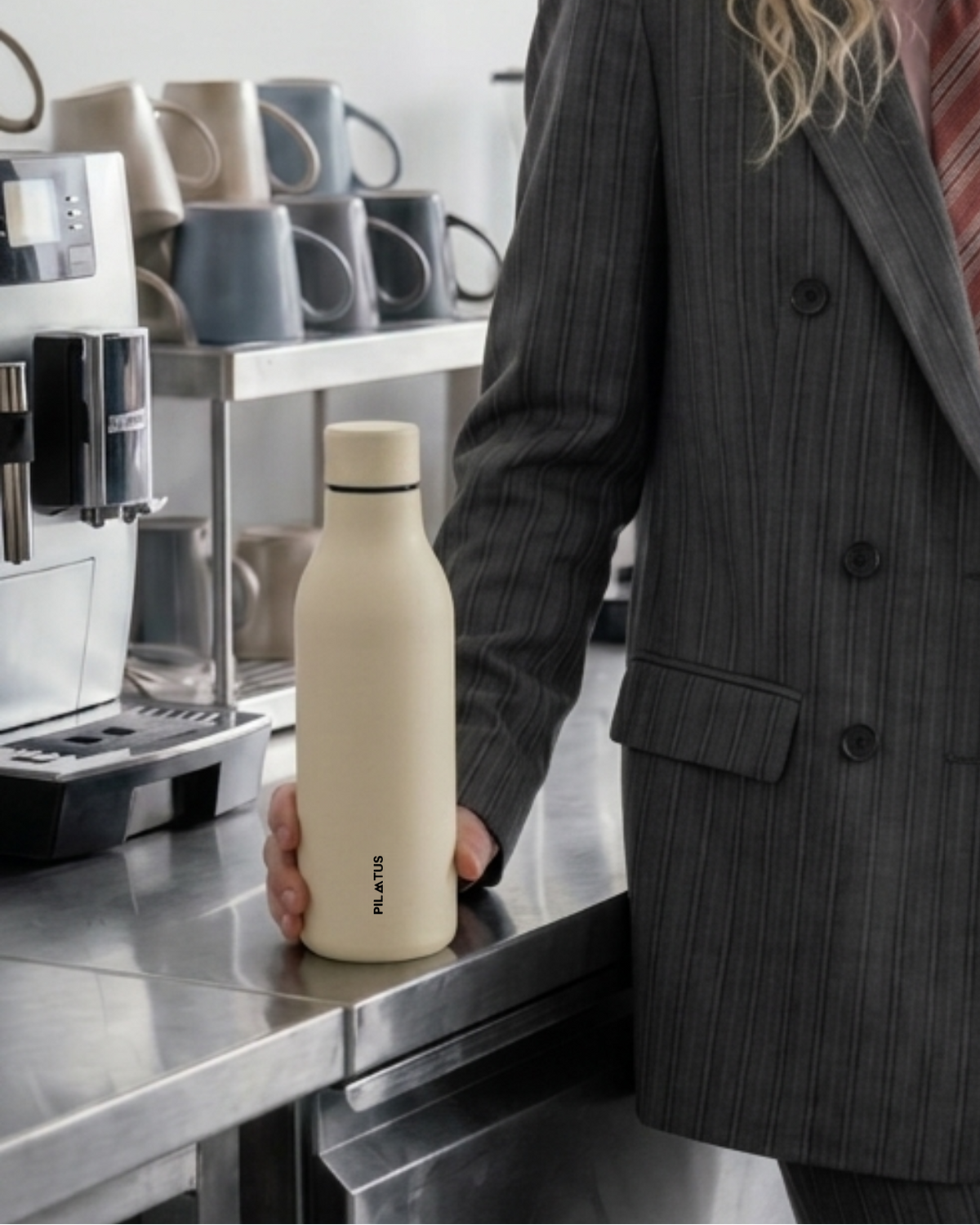 Botella térmica beige con texto vertical PILATUS sostenida sobre encimera metálica; mano sujetándola