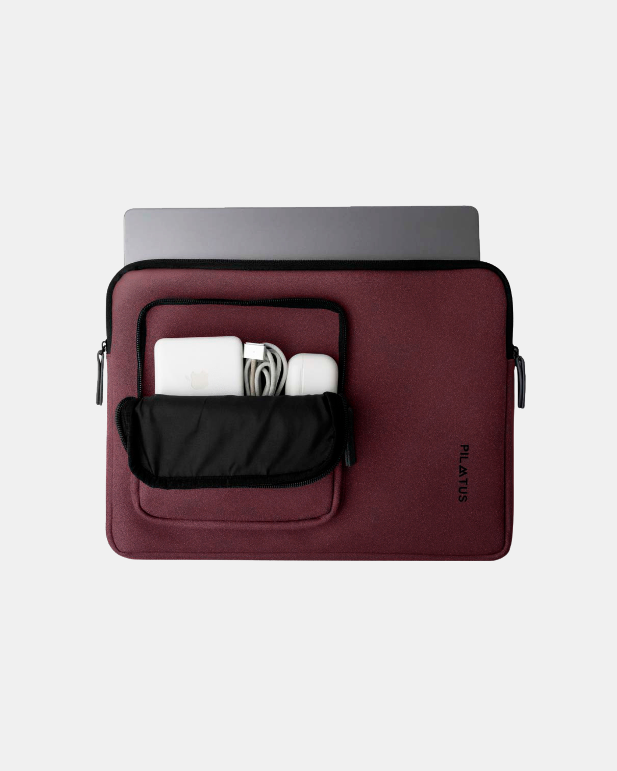 Funda para portátil burdeos con bolsillo frontal abierto que muestra cargador, cable enrollado y auriculares. Texto: PILATUS