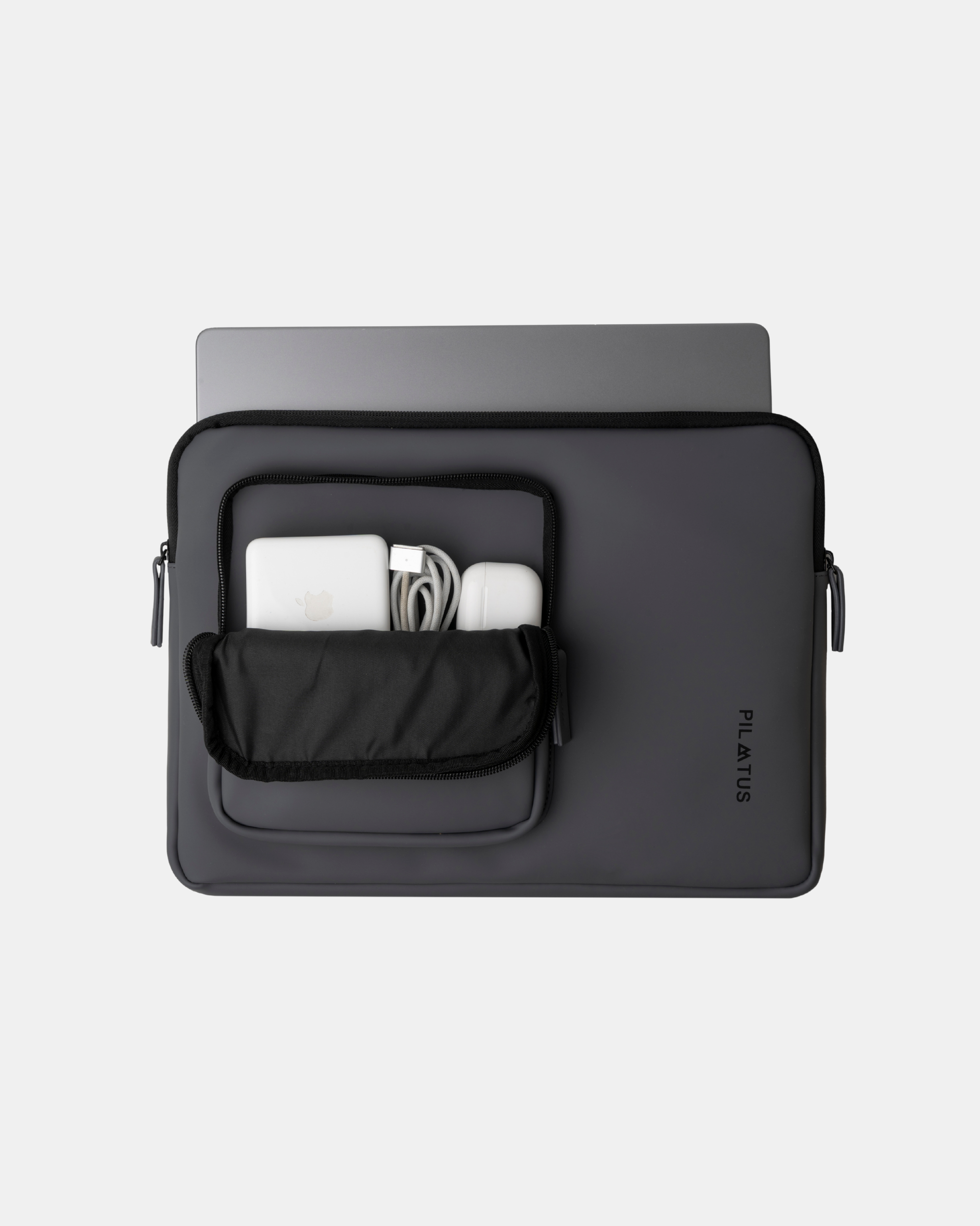 Funda para portátil gris oscuro con bolsillo frontal abierto que muestra cargador, cable y auriculares. Texto: PILATUS