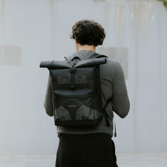 Mochila roll-top negra con logo Red Bull y texto PILATUS en bolsillo frontal; hombre de espaldas llevándola