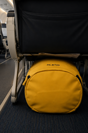 Bolsa circular amarilla colocada bajo el asiento de un avión; alfombra azul y respaldo negro visibles. Texto: PILATUS
