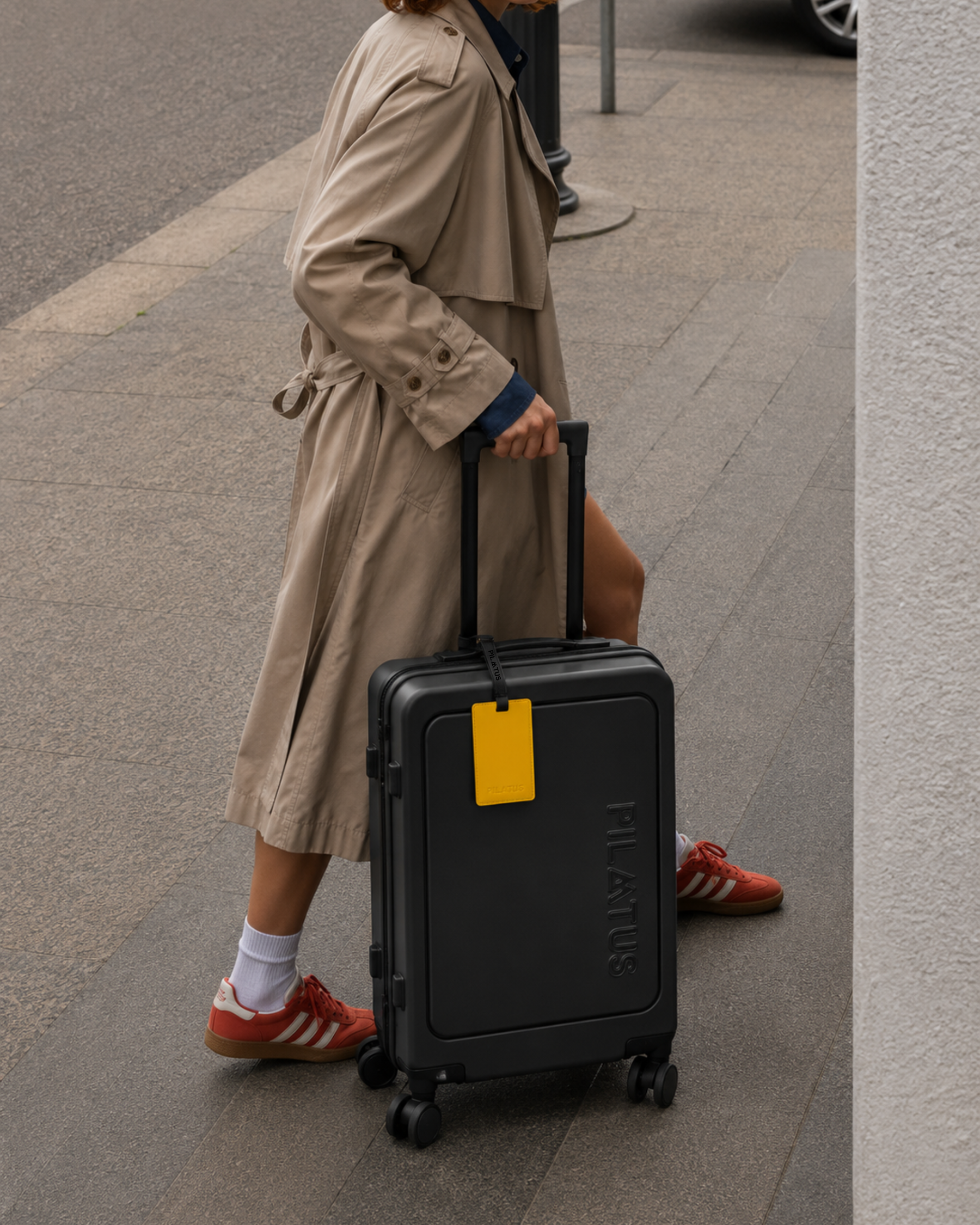 Maleta rígida negra con asa telescópica y etiqueta amarilla; persona con gabardina beige y zapatillas rojas tirando