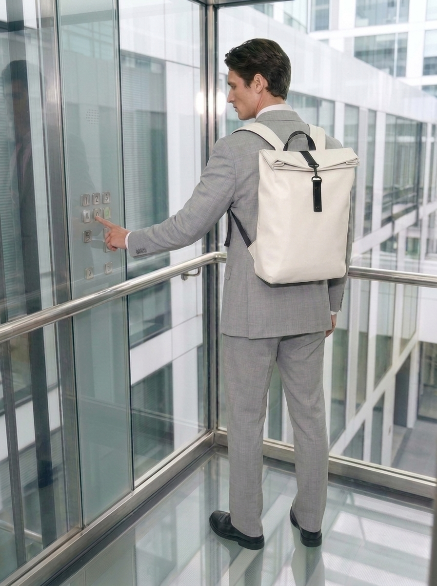 Mochila beige con cierre enrollable y detalle negro; hombre con traje gris en ascensor de cristal en edificio de oficinas